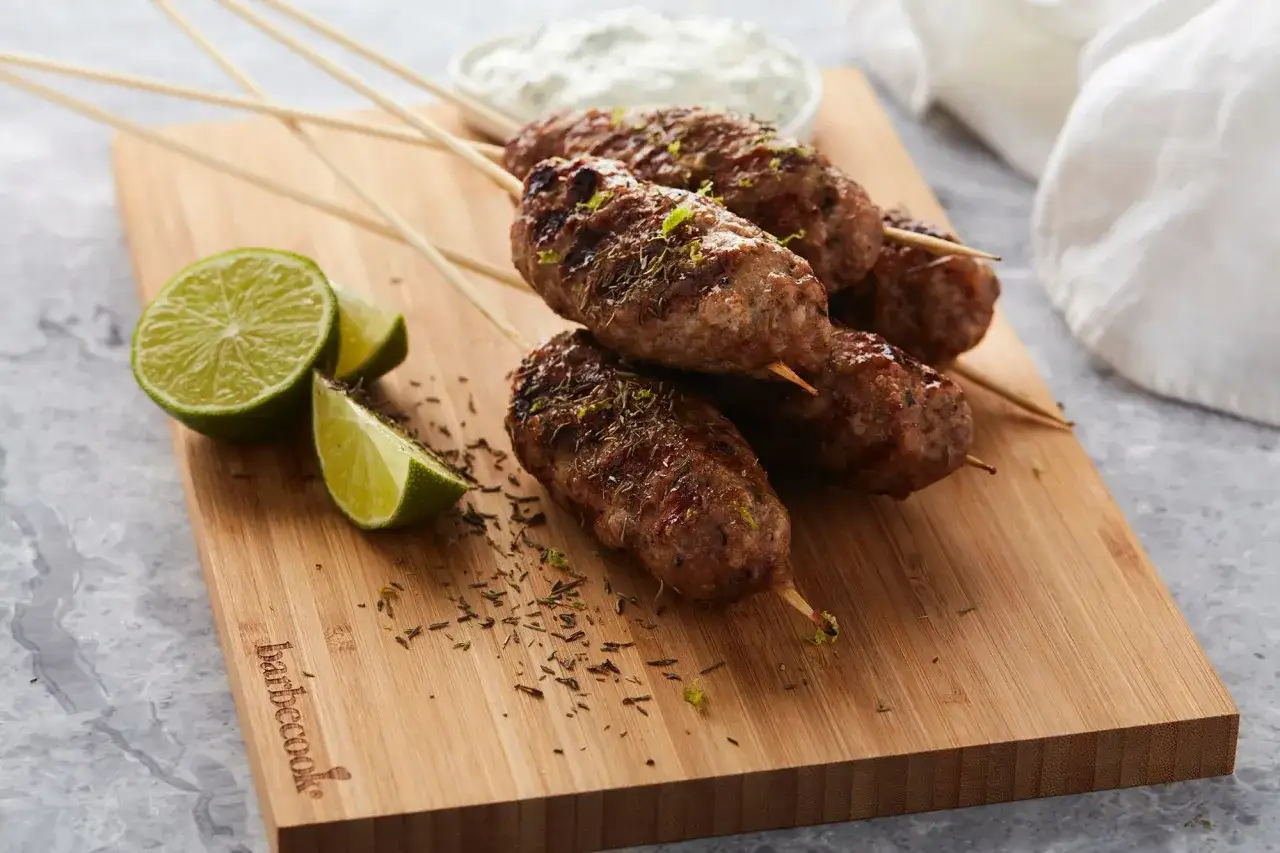 Brochettes d'agneau grillées au barbecue, garnies d'herbes et de zeste de citron vert, servies avec des quartiers de citron vert et une sauce blanche.