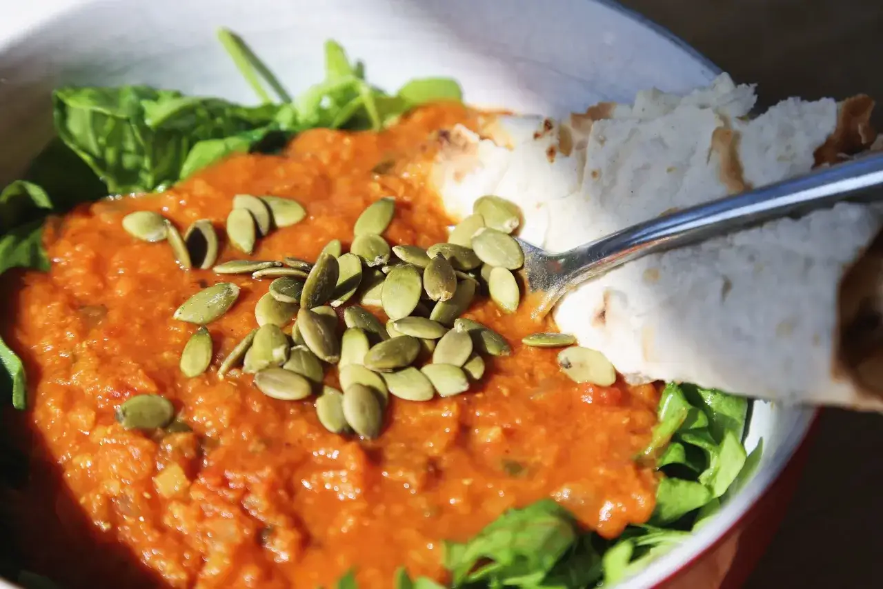 Un bol de pur&eacute;e de lentilles corail, garni de graines de courge et de feuilles vertes, avec un morceau de pain pita.