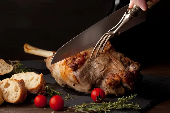 Une main utilise un couteau et une fourchette pour découper un gigot d'agneau rôti, prêt à être servi avec du pain et des tomates.